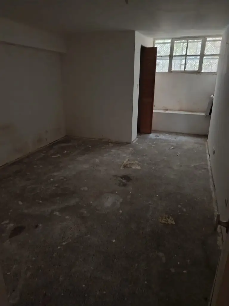 PduF apres - Débarras de Maison Dim, empty room with bare walls, a partial partition, and a row of windows at the far end; dusty concrete floor with debris.