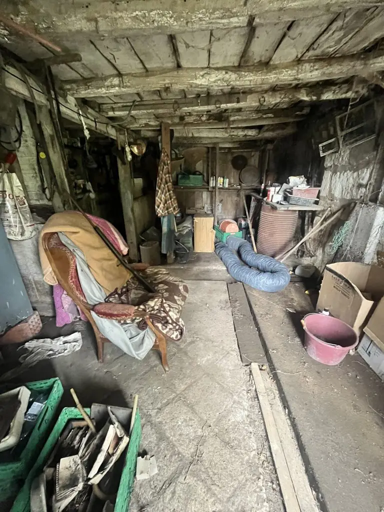 gange avant - Débarras de Maison Dusty, cluttered basement workshop with exposed wooden beams, hoses on the floor, and scattered tools and boxes.